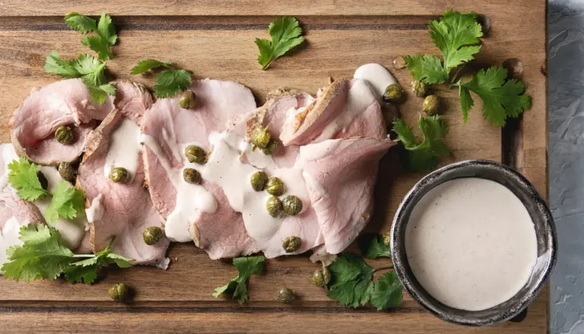 Fette di arrosto con salsa tonnata, capperi e prezzemolo su tagliere di legno.