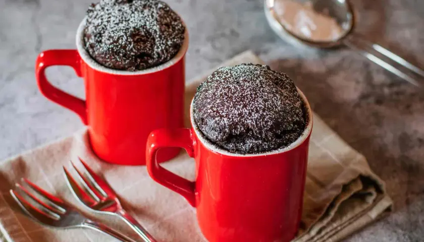 Due tortine in tazza al microonde, ricoperte di zucchero a velo, pronte per essere gustate con due forchettine.
