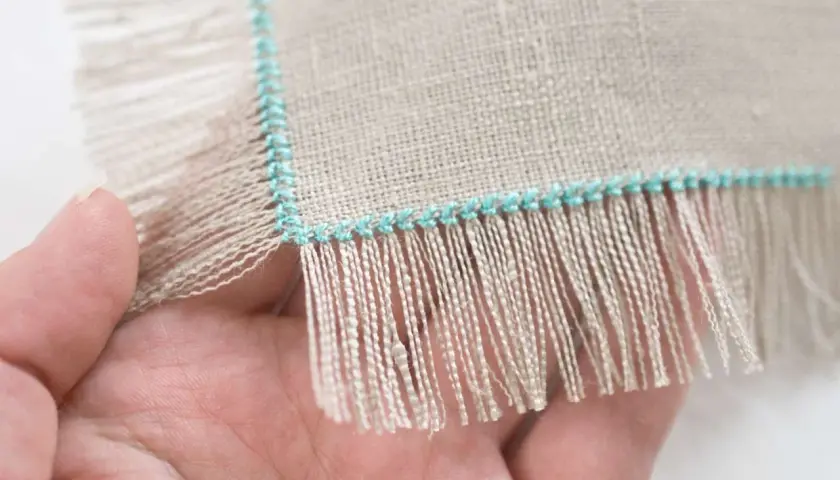 Close-up of a hand holding linen fabric with a turquoise stitched edge, showing how to sew fringe.
