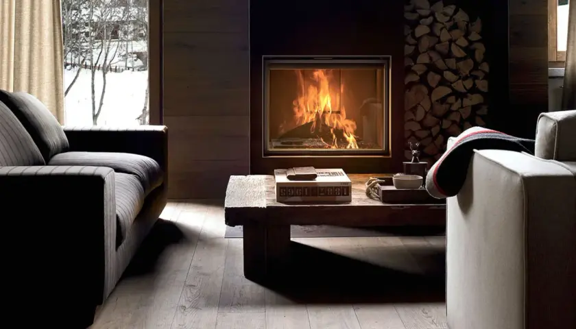 Gemütliches Wohnzimmer mit Kaminfeuer, Holzstapel und Blick auf verschneite Bäume. Tolle Kamin Ideen für Ihr Wohnzimmer.