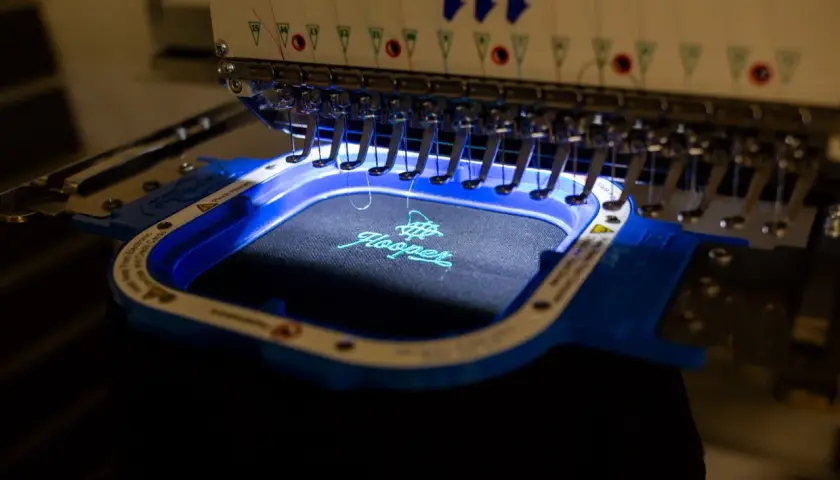 Machine à broder Tajima créant une estampille digitalisée sur un tissu noir. Le logo "Hooper" est en cours de réalisation avec du fil turquoise.