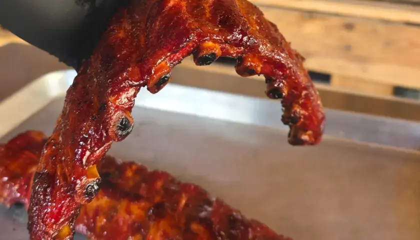 En hand i svart handske håller upp saftiga, grillade revben. De glänser av glaze och ser otroligt goda ut.
