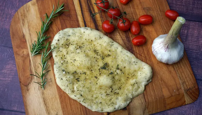Ein unfertiges Pizzabrot mit Knoblauch und Kräutern liegt auf einem Holzbrett, daneben Kirschtomaten und Rosmarin.