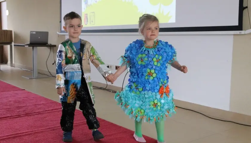 Dzieci prezentują stroje ekologiczne. Dziewczynka w niebieskiej sukience z nakrętek, chłopiec w kurtce z opakowań. Inspiracja, jak zrobić strój ekologiczny dla dziewczynki.