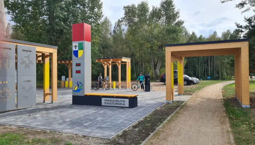 Nowiutka stacja rowerowa na Marszrucie Kaszubskiej. Dwie panie z rowerami, nowoczesna infrastruktura i leśne otoczenie.