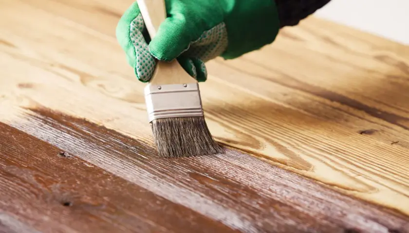 Mano con guante verde aplicando barniz sobre madera, creando un acabado brillante. Barnizar sobre barniz sin lijar para renovar la superficie.