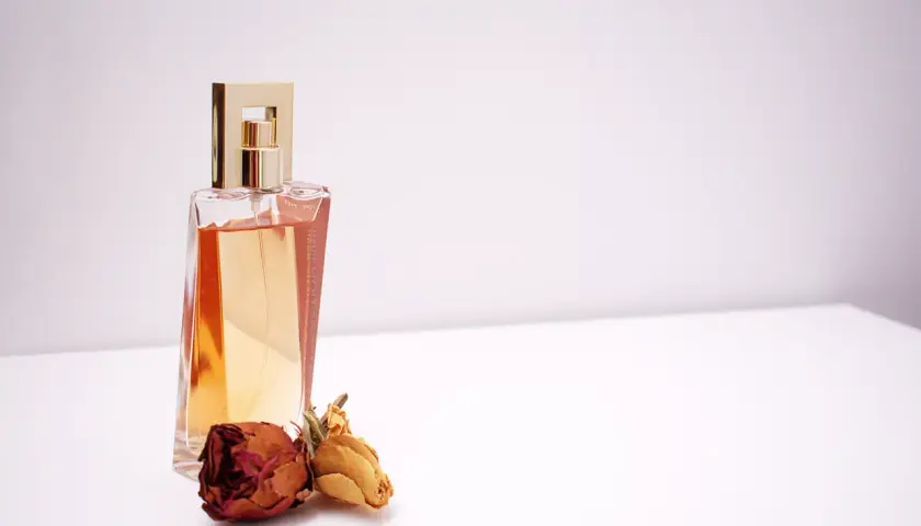 Elegancka butelka perfum z płynem w kolorze bursztynu i złotą nakrętką, obok dwie zasuszone róże. Inspiracja do tworzenia własnych perfum.