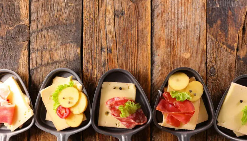 Petites coupelles pour un apéritif raclette, garnies de fromage, charcuterie et pommes de terre sur fond de bois.