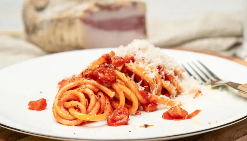 Ein Teller Pasta all'Amatriciana, garniert mit geriebenem Parmesan. Im Hintergrund ein Stück Guanciale, die Zutat für das authentische Amatriciana Rezept.
