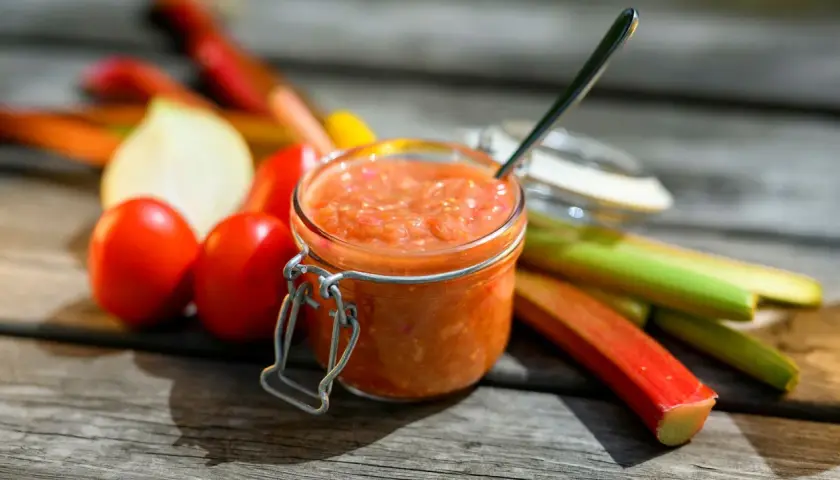 En burk med rabarber chutney står på ett träbord, omgiven av färska rabarber, tomater och en lök.
