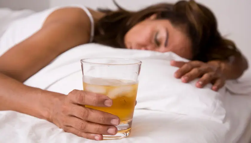 Kobieta śpi w łóżku, trzymając szklankę z napojem i lodem. Może to być alkohol, np. GHB.