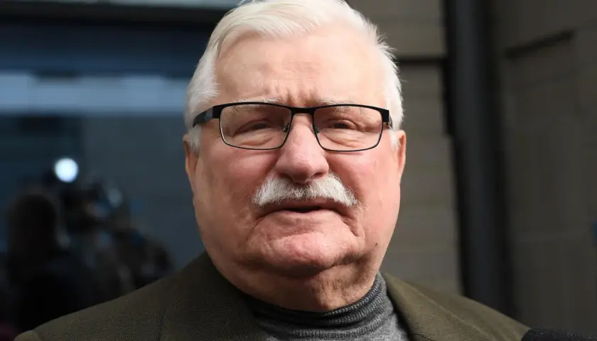 Lech Wałęsa w okularach i z wąsami, w swetrze i marynarce. W tle niewyraźne postacie.