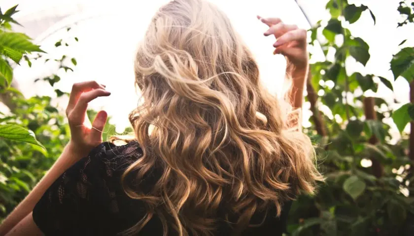Blondes Haar mit Beach Waves im Sonnenlicht, umgeben von grünen Blättern.
