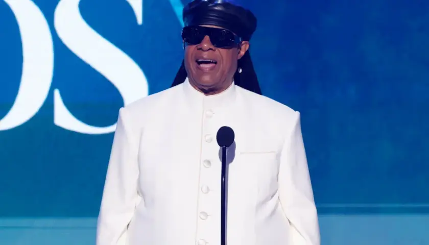Niewidomy piosenkarz Stevie Wonder w białym garniturze i czarnej czapce, przemawia do mikrofonu na tle niebieskiej sceny z logo CBS.