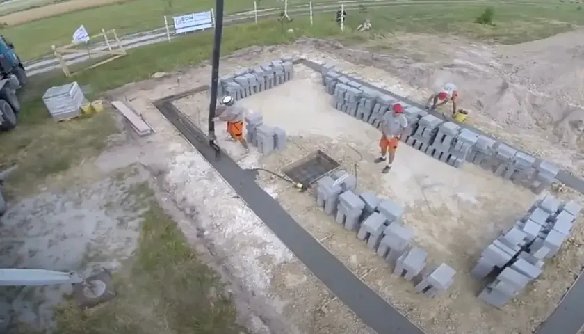 Pracownicy wylewają beton na fundamenty. Widać koparkę, betoniarkę i sterty pustaków.