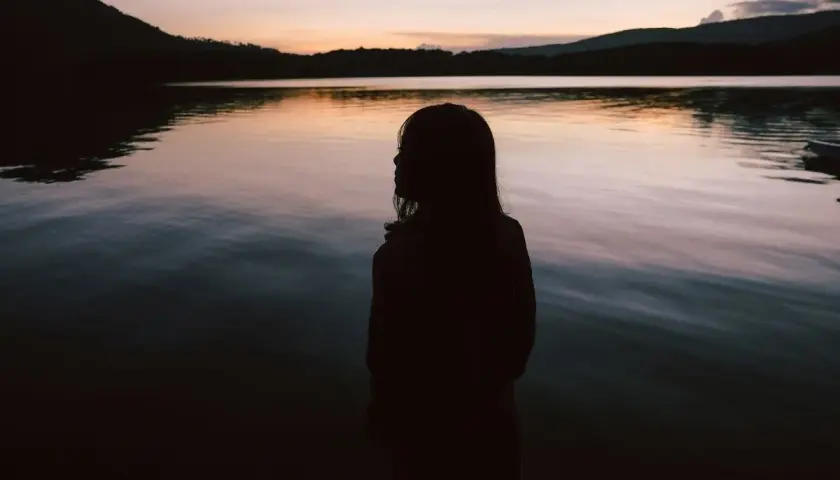 Sylwetka dziewczynki nad jeziorem o zachodzie słońca. Woda odbija niebo, tworząc spokojny obraz. Idealne miejsce na chwilę refleksji, jak w cytaty o spokoju i ciszy.