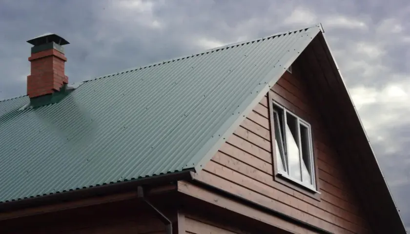 Dach domu pokryty zieloną blachą trapezową, widoczny komin i okno. Szerokość krycia blachy trapezowej zapewnia szczelność.