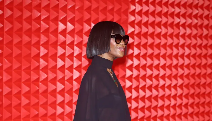 Naomi Campbell luce un corte de pelo **blunt cut** y gafas de sol, posando ante un fondo rojo texturizado.