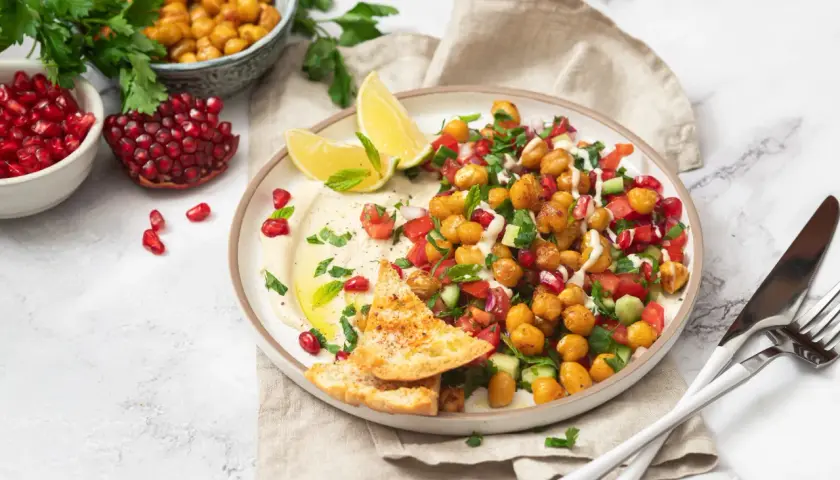 Bunter Kichererbsen Salat mit Granatapfelkernen, Tomaten, Gurken und Hummus, garniert mit Minze und serviert mit Pitabrot.