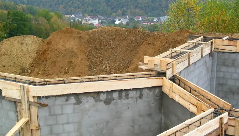 Fundament budowy z szalunkiem i zbrojeniem, w tle domy i las.