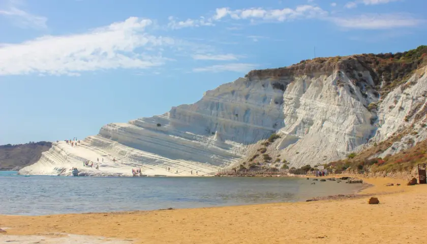 La Scala dei Turchi, une falaise blanche spectaculaire en Sicile, se dresse au-dessus d'une plage de sable doré et d'une mer bleue. Des visiteurs explorent ses terrasses naturelles.