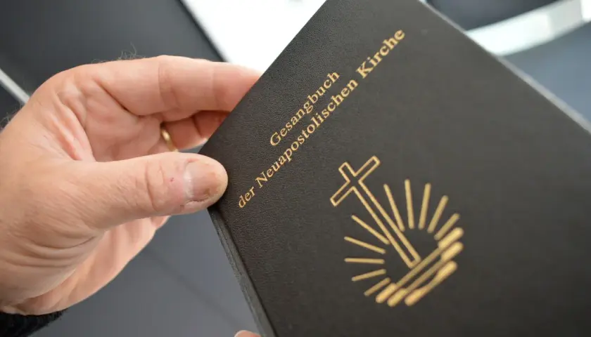 Ein Buch mit dem Titel "Gesangbuch der Neuapostolischen Kirche" wird von einer Hand gehalten. Das Symbol der Kirche, ein Kreuz mit Strahlen, ist auf dem Einband abgebildet.