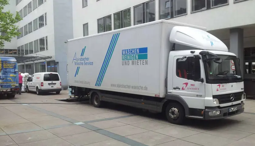 Ein LKW von Alpirsbacher Wäsche Service, der für Werkverkehr genutzt wird, steht vor einem Gebäude. Er transportiert Wäsche.
