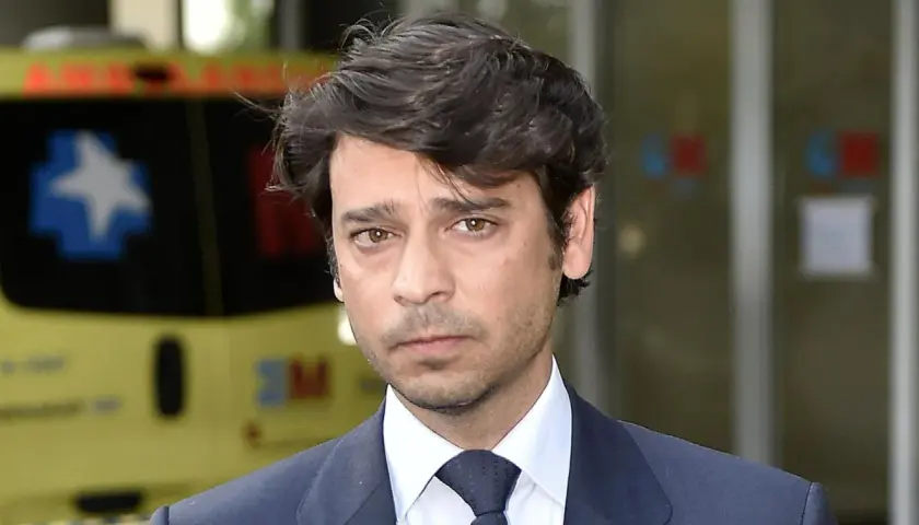 Miguel Palomo Danko, con traje oscuro y corbata, mira fijamente. Detrás, una ambulancia amarilla.