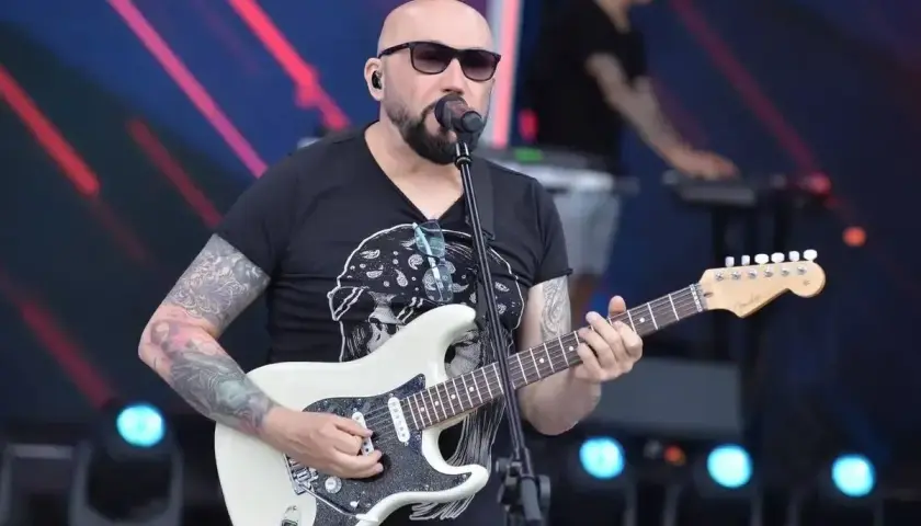 Artysta z tatuażami gra na białej gitarze, śpiewając do mikrofonu. W tle widać klawiszowca. To z pewnością niezapomniane **kombi utwory** na żywo!
