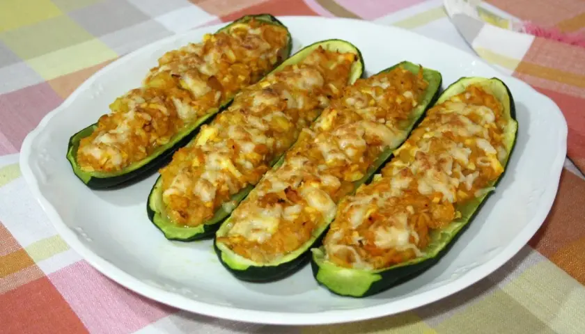 Cuatro calabacines rellenos de atún, gratinados con queso, servidos en un plato blanco sobre un mantel a cuadros.