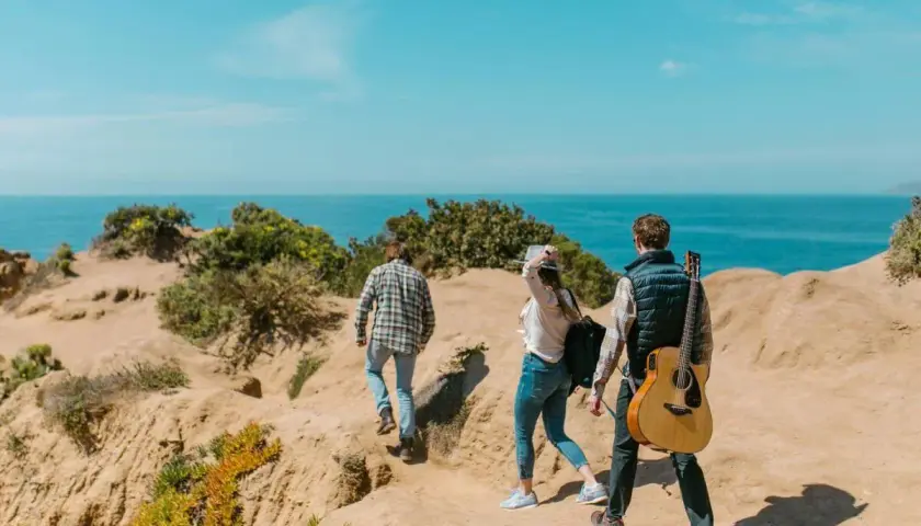 Trzy osoby wędrują ścieżką nad morzem. Jeden z nich niesie gitarę. Idealne szlaki dla dzieci na wakacyjną przygodę.