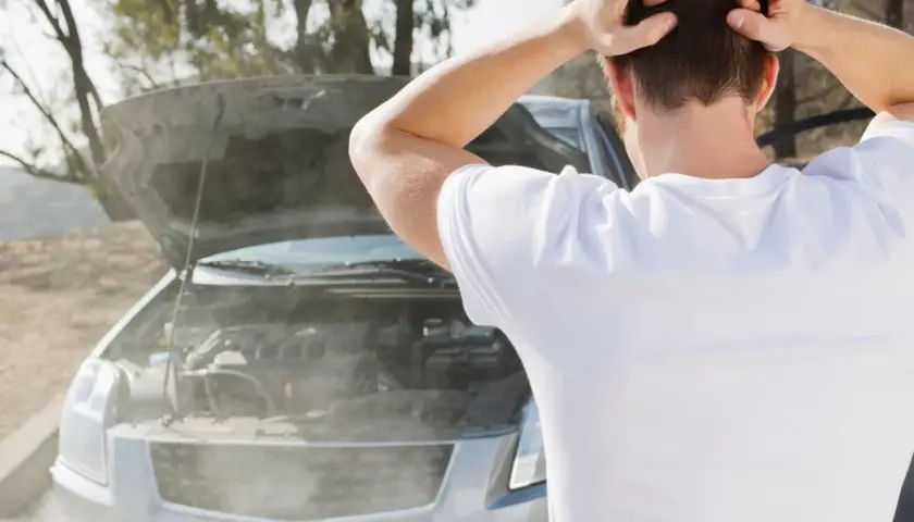 Hombre frustrado mira cómo sale humo blanco del motor de su coche averiado.