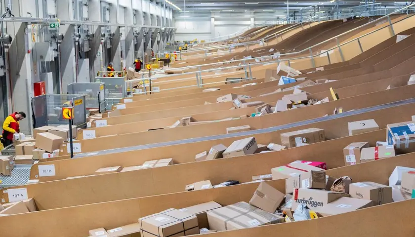 Arbeiter sortieren Pakete in einem riesigen DHL-Zentrallager. Zahlreiche Pakete liegen auf Förderbändern und in Regalen.