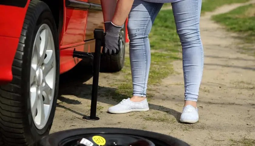 Kobieta wymienia koło w czerwonym samochodzie. Obok leży opona zapasowa z żółtą naklejką "80". Zastanawiasz się, ile można jeździć na oponie zapasowej?