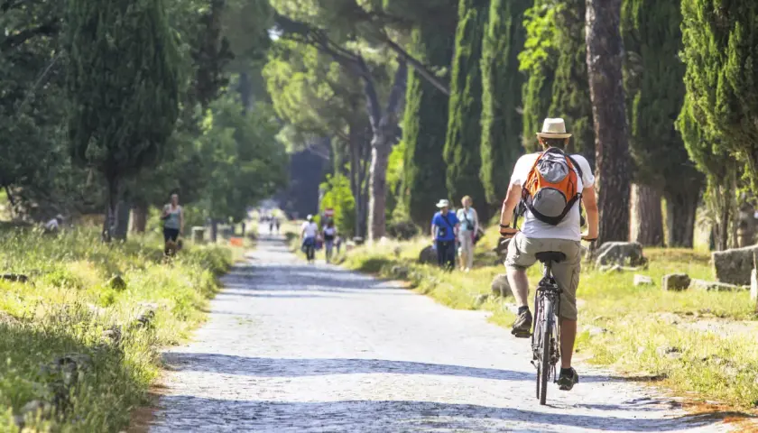 Un hombre pedalea por un camino empedrado en Roma, rodeado de altos cipreses y ruinas antiguas.