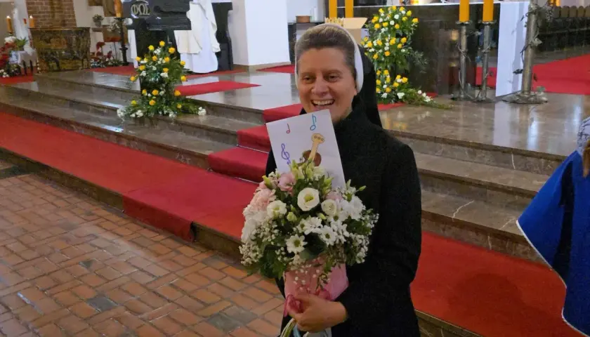 Radosna zakonnica z bukietem kwiatów i kartką z nutami. W tle msza święta, 25 października.