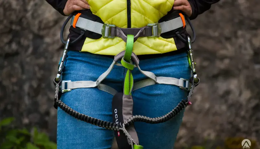 Osoba w kamizelce i jeansach ma na sobie sprzęt do via ferrata, w tym lonżę z karabinkami.