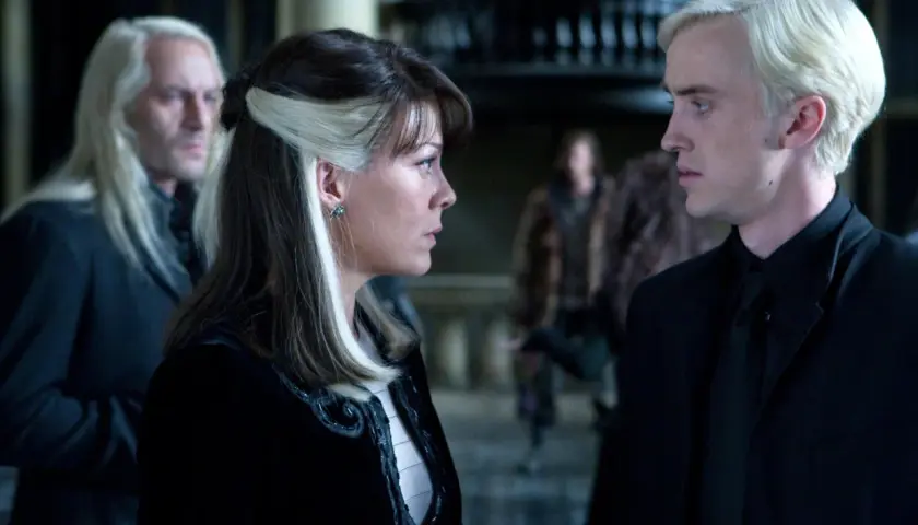 Narcyza Malfoy rozmawia z synem, Draco, w ponurym wnętrzu. W tle widać Lucjusza Malfoya.