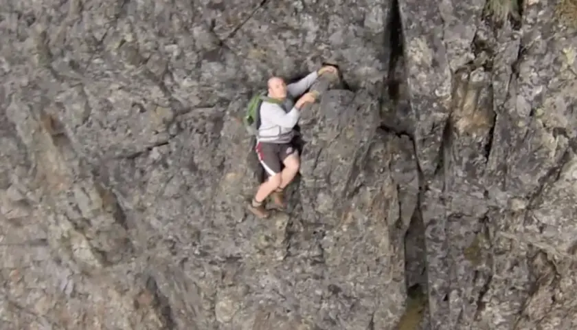 A man is climbing a steep, rocky cliff face. He's wearing shorts and a backpack, looking like he's truly "cliffed out" and facing a challenging ascent.