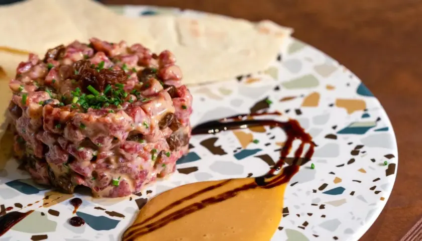 Delicioso tartar de salchichón, con trocitos de frutos secos y cebollino fresco, servido en un plato moderno con salsas.