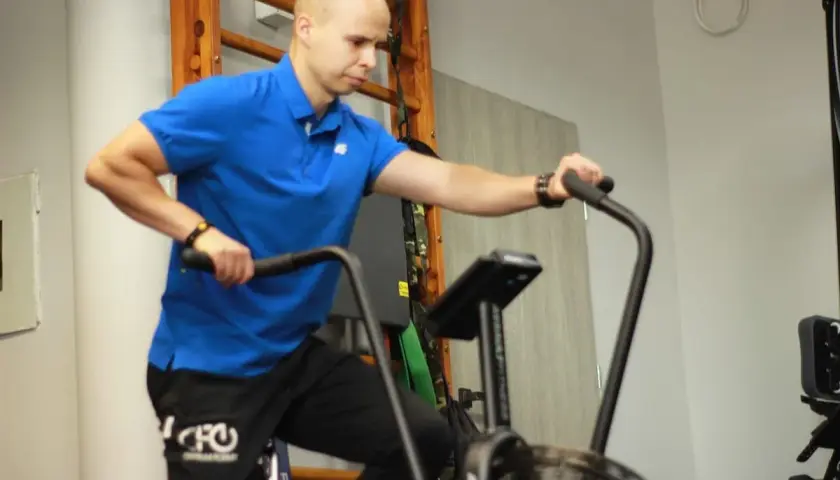 Mężczyzna w niebieskiej koszulce wykonuje trening aerobowy na rowerze stacjonarnym.