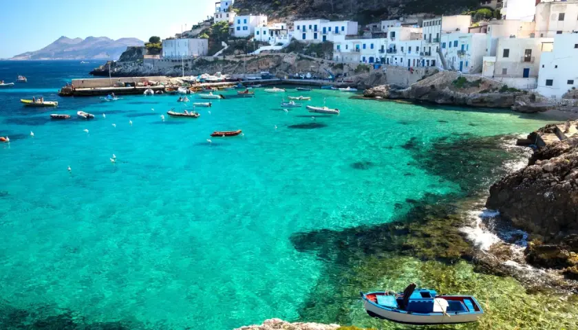 Boats in a turquoise bay near white buildings, a picturesque scene reminiscent of the charm found near international airports in Sicily.