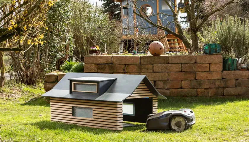 Ein moderner, selbstgebauter Rasenroboter-Unterstand mit Holzlamellen und Dachfenstern. Der Rasenroboter parkt daneben.