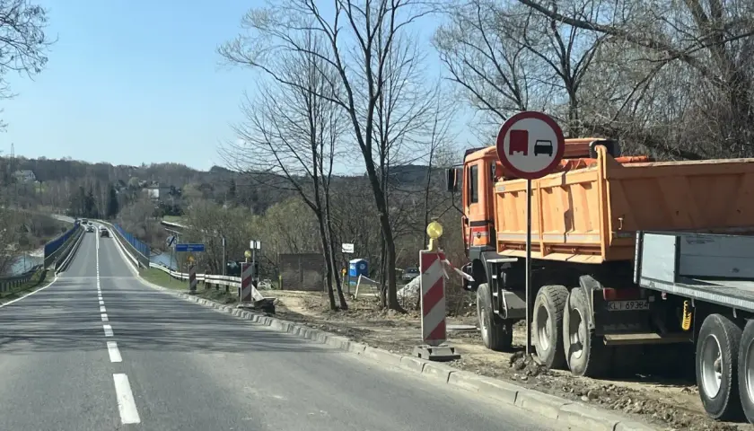 Pomarańczowa wywrotka MAN stoi przy drodze, gdzie trwa remont. Znak zakazu wyprzedzania dla pojazdów ciężarowych. Velo Raba w tle.