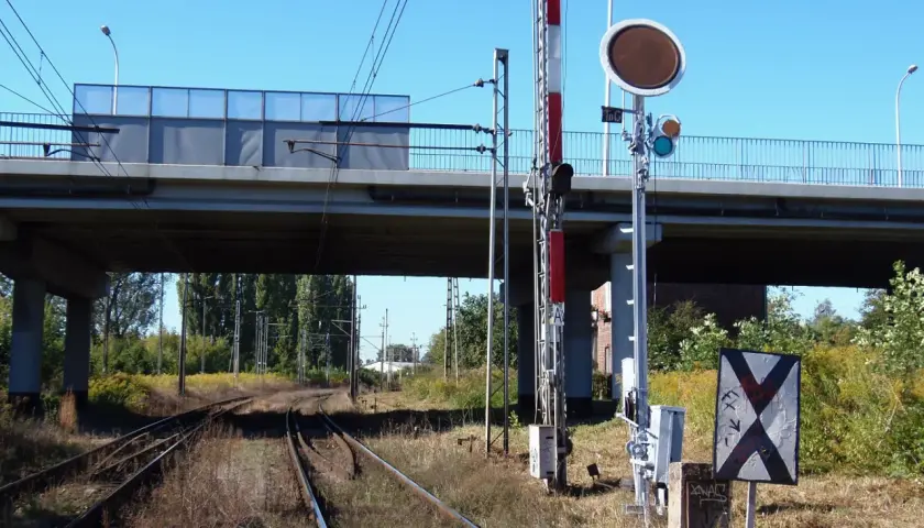 Tarcza ostrzegawcza przy torach kolejowych pod wiaduktem. Sygnalizacja świetlna i znak krzyżowy informują o zbliżającym się niebezpieczeństwie.