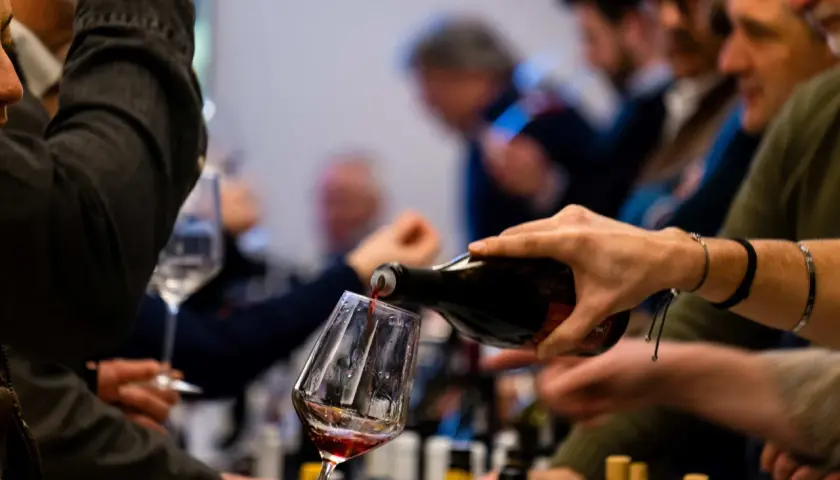 Versando vino rosso in un calice, si apprezzano le caratteristiche del vermentino. Persone in attesa, bottiglie sullo sfondo.