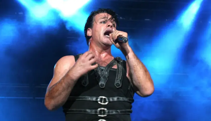 Till Lindemann na koncercie, w czarnym, skórzanym stroju, śpiewa do mikrofonu na tle niebieskich świateł.