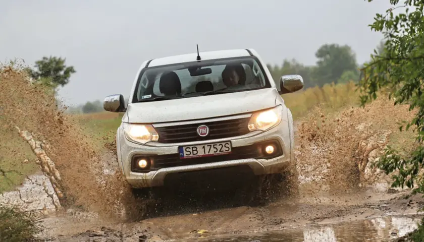 Biały Fiat Fullback z impetem wjeżdża w błotnistą kałużę, wzbijając fontanny brudu.