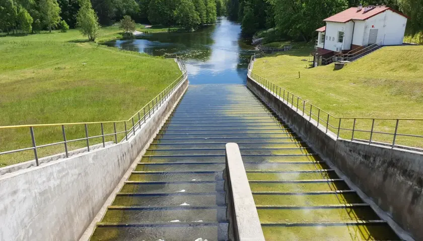 Widok na stopnie wodne zapory Mylf, z zielonymi trawnikami i budynkiem technicznym po prawej.