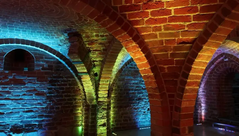 Kamienne łuki i ceglane ściany tworzą tajemniczą atmosferę. Piwnica romańska oświetlona jest kolorowymi światłami, tworząc magiczny nastrój.